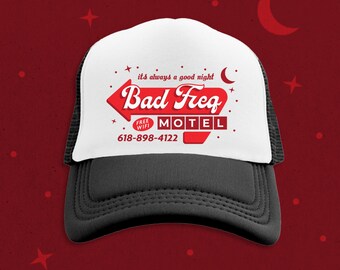 May include: A white and black trucker hat with a red and white motel sign graphic. The sign says "Bad Freq Motel" with the text "it's always a good night" above it. The sign also has a small red square with the text "Free Wifi" inside. The phone number 618-898-4122 is printed below the sign.