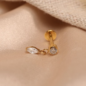 Può includere: Un orecchino dorato con una piccola gemma rotonda e trasparente e una gemma trasparente a forma di marquise pendente. L'orecchino è posto su un tessuto color pesca.