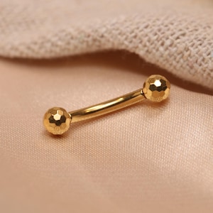 May include: A curved, gold-colored barbell body piercing. The ends of the barbell are spherical with a faceted, textured surface. The barbell is displayed on a light pink satin-like fabric.