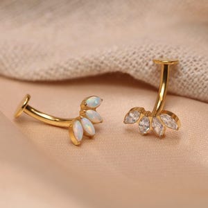 Puede incluir: Dos piercings dorados para el ombligo. Uno presenta tres piedras tipo ópalo, y el otro tiene tres piedras transparentes con forma de diamante. Las joyas se exhiben sobre una tela color melocotón.