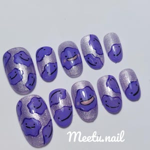 May include: A set of ten artificial nails with a purple and silver glitter design. Each nail features a cartoon character with a smiling face. The text "Meetu.nail" is written in white.