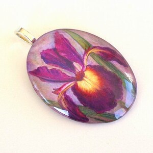 May include: An oval-shaped pendant featuring a detailed illustration of a deep purple iris flower with a bright yellow center and green leaves. The pendant has a smooth, glossy finish and includes a silver-toned metal bail at the top.