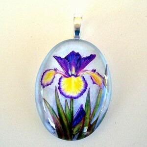 May include: Oval pendant featuring a detailed painting of a purple and yellow iris flower. The pendant has a silver-colored bail and is encased in clear resin. The flower's petals and leaves are visible within the transparent setting.