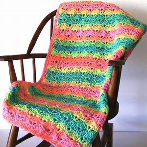 May include: A colorful, handmade crochet blanket in a rainbow of hues, including pink, orange, yellow, green, and blue. The blanket is draped over a wooden rocking chair, showcasing its textured, shell-like stitch pattern. A cozy, vibrant home decor item.