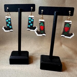 May include: Two pairs of beaded earrings shaped like cowboy boots. One pair features blue floral designs, while the other has a red strawberry design. Both pairs have white and black detailing and gold-colored ear wires.