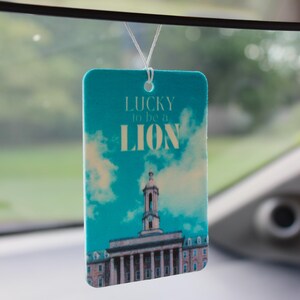 Op de afbeelding: Rechthoekige luchtverfrisser met een turquoise achtergrond met wolken en een gebouw. De tekst "LUCKY to be a LION" is in het wit gedrukt. Een wit touwtje is bevestigd om op te hangen.