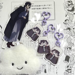 Peut inclure: Un ensemble de quatre porte-clés de personnages d'anime représentant un personnage masculin aux cheveux noirs et un manteau violet. Les porte-clés sont attachés à un charme en forme de cœur violet avec un anneau métallique. Les porte-clés sont présentés sur une page d'un livre avec du texte japonais.