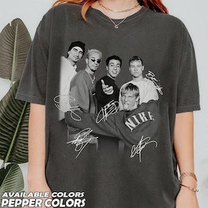 Pode incluir: Uma t-shirt de manga curta cinzento-carvão com uma impressão gráfica a preto e branco da boy band Backstreet Boys. O design inclui os cinco membros com as suas assinaturas impressas na frente, um dos quais usa uma camisola Nike.