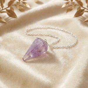 Könnte beinhalten: Ein facettierter, kegelförmiger Amethyst-Kristallanhänger an einer feinen silberfarbenen Metallkette. Der violette Edelstein weist natürliche Einschlüsse und eine polierte Oberfläche auf, was für ein durchscheinendes Aussehen mit Lichtreflexen sorgt.