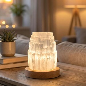 Könnte beinhalten: Eine zylindrische Selenit-Kristalllampe mit einer durchscheinenden, geschichteten und faserigen Textur. Die Lampe wird von innen beleuchtet, strahlt ein sanftes weißes Licht aus und ist auf einem runden, hellen Holzsockel mit Stromkabel montiert.