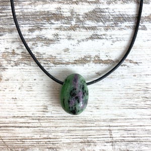 Puede incluir: Un collar con colgante de piedra preciosa verde y morada. La piedra de forma ovalada está suspendida de un cordón negro. La piedra tiene un patrón moteado de tonos verdes y morados. El fondo es una superficie de madera clara.