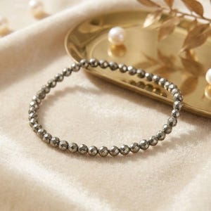 Peut inclure: Un bracelet de perles composé de petites pierres de pyrite grises et facettées. Le bracelet est circulaire et repose sur une surface veloutée de couleur crème. Un plateau doré avec des perles et des feuilles décoratives est en arrière-plan.