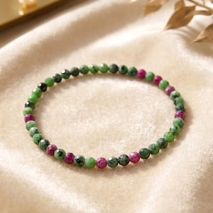 Könnte beinhalten: Ein kreisförmiges Perlenarmband mit kleinen, facettierten, runden Perlen in abwechselnden Farbtönen von leuchtendem Grün und tiefem Magenta. Die Perlen sind zu einer durchgehenden Schlaufe aufgereiht und weisen eine polierte Textur auf.