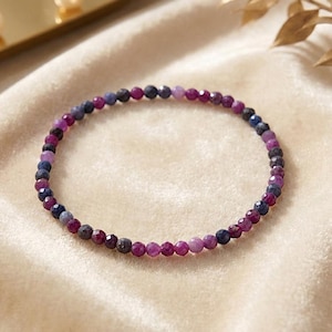 Könnte beinhalten: Ein zartes Perlenarmband mit kleinen, facettierten runden Perlen in abwechselnden Farbtönen von tiefem Magenta, Dunkelblau und hellem Violett. Die Perlen sind in einem durchgehenden kreisförmigen Muster aufgereiht und bilden ein schmales Design.