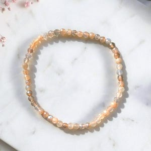 Könnte beinhalten: Ein zartes Stretch-Armband bestehend aus kleinen, facettierten, runden Perlen in verschiedenen Nuancen von Pfirsich, Beige und durchscheinendem Grau. Die Perlen haben eine subtil reflektierende Oberfläche, die einen sanften Schimmer erzeugt.