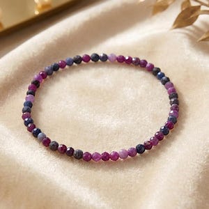 Könnte beinhalten: Ein zartes Perlenarmband mit kleinen, facettierten runden Perlen in abwechselnden Farbtönen von tiefem Magenta, Dunkelblau und hellem Violett. Die Perlen sind in einem durchgehenden kreisförmigen Muster aufgereiht und bilden ein schmales Design.