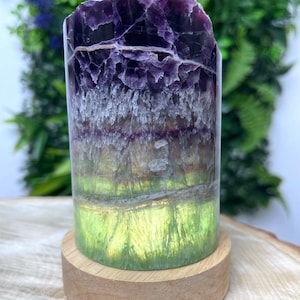 Könnte beinhalten: Zylindrischer Fluoridkristall mit Schichten in Lila, Grau und Grün. Der Kristall steht auf einem runden Holzsockel, der seine natürlichen Farben und Texturen hervorhebt. Die Oberseite des Kristalls hat eine raue, unebene Oberfläche.
