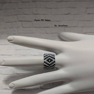 May include: A beaded ring in black and white, featuring a geometric pattern. The ring is displayed on a white hand against a white brick background. The text "Peyote PDF Pattern" and "By: JerzeyGems" are visible.