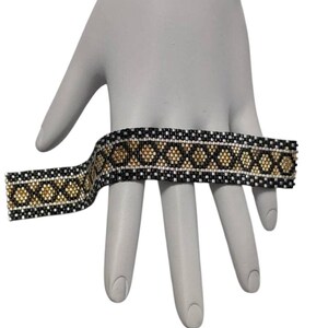 May include: A wide beaded bracelet featuring a repeating geometric pattern of black, metallic gold, and silver seed beads. The design consists of a central row of gold diamond shapes bordered by black and silver horizontal lines in a woven style.