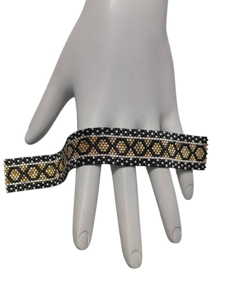 May include: A wide beaded bracelet featuring a repeating geometric pattern of black, metallic gold, and silver seed beads. The design consists of a central row of gold diamond shapes bordered by black and silver horizontal lines in a woven style.