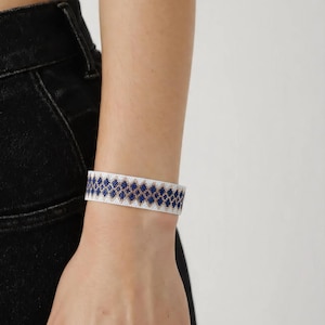 May include: A wide cuff bracelet featuring a repeating geometric diamond pattern in deep blue and metallic gold, set against a white background. The bracelet is constructed from small, uniform beads arranged in a structured, flat band design.