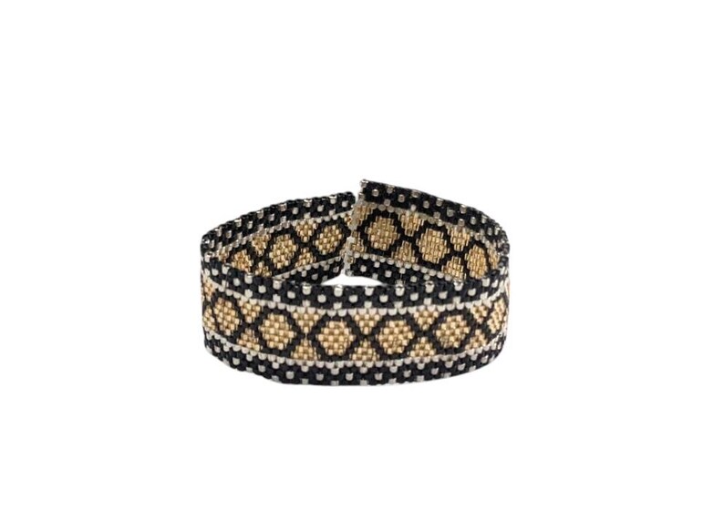 May include: A woven beaded bracelet featuring a repeating geometric diamond pattern in black, metallic gold, and white. The band is constructed from small, uniform beads arranged in horizontal rows, creating a textured, flexible, and circular structure.