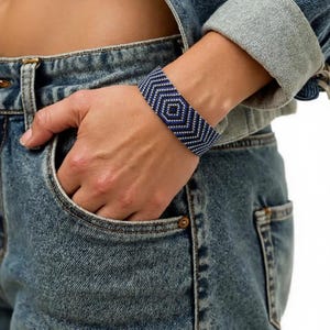 May include: A wide woven bracelet featuring a geometric chevron pattern in alternating rows of deep blue and metallic silver beads. The bracelet has a structured, flat band design that wraps around the wrist, showcasing a repeating diamond motif.