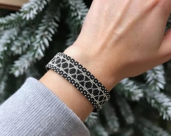 Könnte beinhalten: Ein breites Armband mit einem sich wiederholenden geometrischen Muster aus kleinen schwarzen und silbernen Perlen. Das Design besteht aus einer zentralen Reihe ineinandergreifender Rauten, die von geraden Linien aus silbernen Perlen auf schwarzem Grund gesäumt sind.
