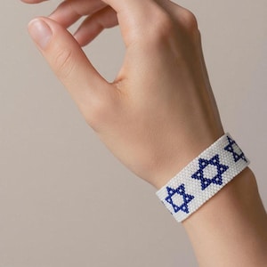Op de afbeelding: Een witte kralenarmband met een herhalend patroon van blauwe Davidssterren. De armband is gemaakt van kleine, uniforme kralen die in een platte, brede band zijn gerangschikt die om de pols sluit, met een duidelijk contrast tussen de blauwe sterren en de witte achtergrond.
