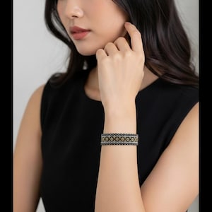 May include: A wide cuff bracelet featuring a repeating geometric pattern in black, gold, and silver tones. The design consists of a central row of gold-colored circular motifs bordered by thin silver lines and black edges with a textured appearance.