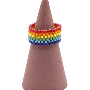 May include: A beaded ring in a rainbow color pattern, displayed on a brown cone-shaped stand. The ring features red, orange, yellow, green, blue, and purple beads. The stand is a solid, muted brown color.