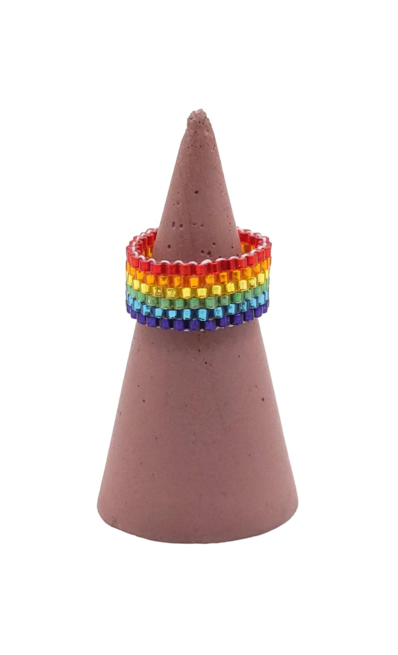 May include: A beaded ring in a rainbow color pattern, displayed on a brown cone-shaped stand. The ring features red, orange, yellow, green, blue, and purple beads. The stand is a solid, muted brown color.