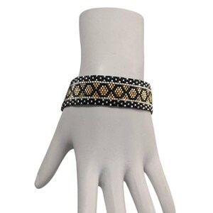 May include: A woven beaded bracelet featuring a geometric pattern of black, white, and metallic gold beads. The design consists of a central row of gold diamond shapes bordered by horizontal lines of black and white beads in a repeating grid structure.