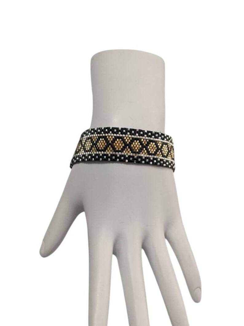 May include: A woven beaded bracelet featuring a geometric pattern of black, white, and metallic gold beads. The design consists of a central row of gold diamond shapes bordered by horizontal lines of black and white beads in a repeating grid structure.
