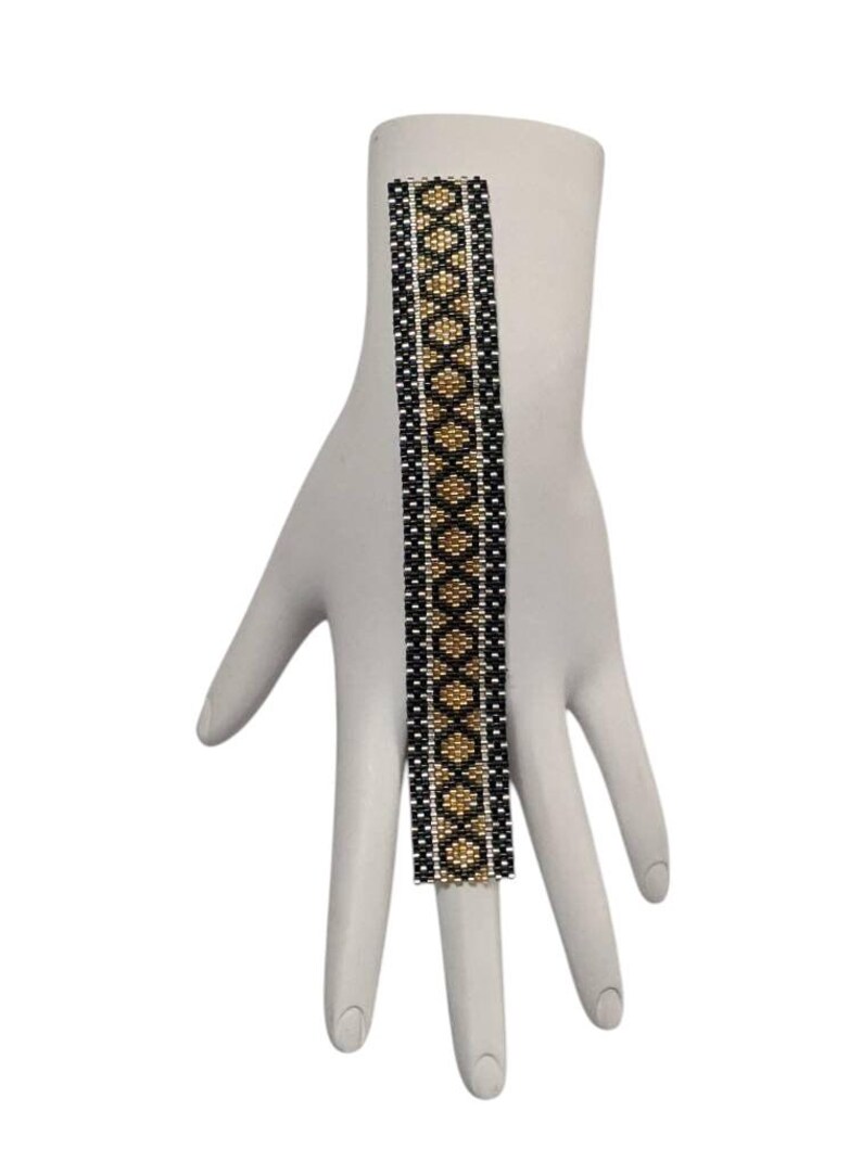 May include: A rectangular beaded bracelet featuring a geometric diamond pattern in black, gold, and silver seed beads. The design consists of a central column of gold diamonds bordered by black and silver vertical stripes, creating a structured look.