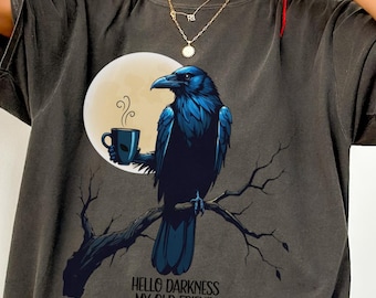 May include: Dark grey t-shirt featuring a blue raven perched on a branch, holding a coffee cup in front of a full moon. The text "HELLO DARKNESS MY OLD FRIEND" is below the raven. The shirt has short sleeves and a relaxed fit.