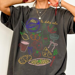 K&ouml;nnte beinhalten: Dunkelgraues T-Shirt mit einer farbenfrohen Grafik italienischer Speisen und Getr&auml;nke, darunter Fisch, Weinflasche, Pasta und K&auml;se. Die Worte "la dolce vita" und "ciao" sind ebenfalls auf dem Shirt zu sehen.