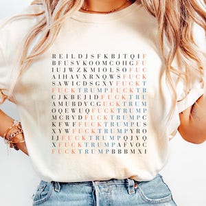 May include: A cream-coloured t-shirt featuring a grid of letters and the phrase "FUCKTRUMP" in red and blue. The shirt is worn with blue jeans. The word "Ivory" is written in cursive in the bottom right corner.