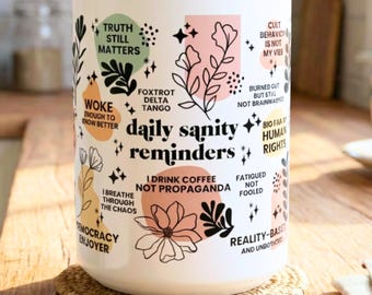 Op de afbeelding: Witte keramische mok met de tekst "daily sanity reminders" en andere zinnen. Het heeft bloemen en geometrische ontwerpen in zwart, perzik en groen. De mok staat op een geweven onderzetter.