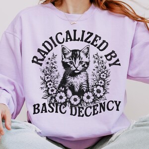 Könnte beinhalten: Ein helllila Sweatshirt mit dem Text "RADICALIZED BY BASIC DECENCY" in Schwarz. Die Grafik zeigt eine schwarz-weiße Katze, umgeben von Blumen. Das Sweatshirt hat einen lässigen Stil.