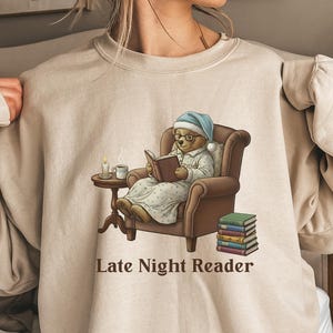 Gildan 18000 sweatshirt featuring a cozy teddy bear wearing a nightgown and hat, sitting in a chair reading a book with a candle, coffee cup, and stack of books beside him. The design includes the text “Late Night Reader” beneath the image. Styled in a warm, vintage-inspired look perfect for book lovers, cozy fall nights, and readers who enjoy a comforting, bookish aesthetic.