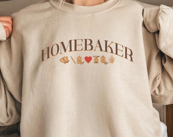 May include: A beige sweatshirt with the text "Homebaker" in brown lettering. The text is arched and has small illustrations of baking tools underneath.