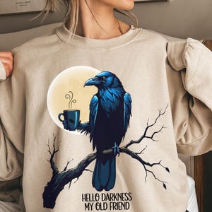 May include: Beige sweatshirt featuring a blue raven perched on a branch, holding a mug of coffee in front of a full moon. The text "HELLO DARKNESS MY OLD FRIEND" is below the raven. The sweatshirt has long sleeves.