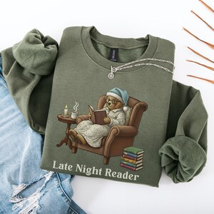 Gildan 18000 sweatshirt featuring a cozy teddy bear wearing a nightgown and hat, sitting in a chair reading a book with a candle, coffee cup, and stack of books beside him. The design includes the text “Late Night Reader” beneath the image. Styled in a warm, vintage-inspired look perfect for book lovers, cozy fall nights, and readers who enjoy a comforting, bookish aesthetic.