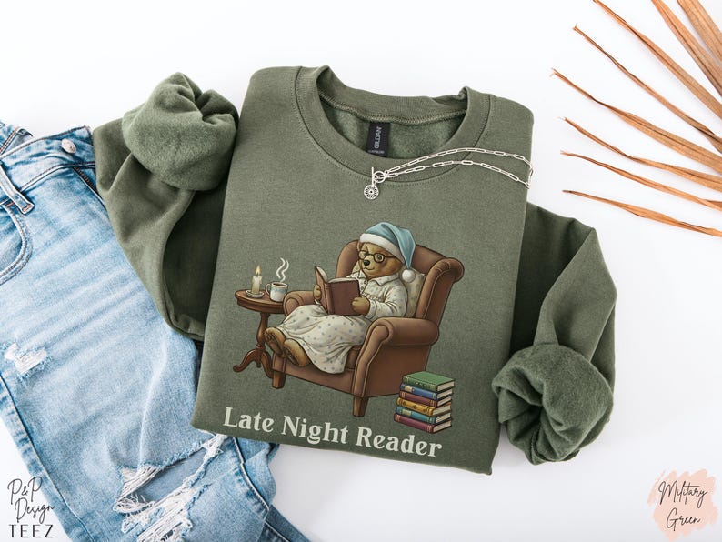 Gildan 18000 sweatshirt featuring a cozy teddy bear wearing a nightgown and hat, sitting in a chair reading a book with a candle, coffee cup, and stack of books beside him. The design includes the text “Late Night Reader” beneath the image. Styled in a warm, vintage-inspired look perfect for book lovers, cozy fall nights, and readers who enjoy a comforting, bookish aesthetic.