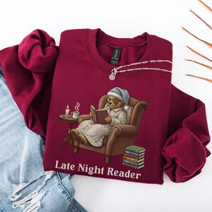 Gildan 18000 sweatshirt featuring a cozy teddy bear wearing a nightgown and hat, sitting in a chair reading a book with a candle, coffee cup, and stack of books beside him. The design includes the text “Late Night Reader” beneath the image. Styled in a warm, vintage-inspired look perfect for book lovers, cozy fall nights, and readers who enjoy a comforting, bookish aesthetic.