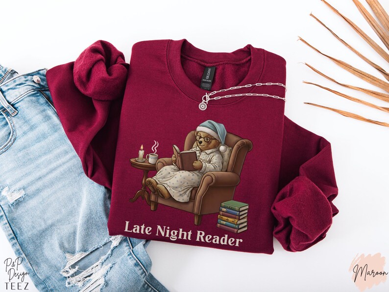 Gildan 18000 sweatshirt featuring a cozy teddy bear wearing a nightgown and hat, sitting in a chair reading a book with a candle, coffee cup, and stack of books beside him. The design includes the text “Late Night Reader” beneath the image. Styled in a warm, vintage-inspired look perfect for book lovers, cozy fall nights, and readers who enjoy a comforting, bookish aesthetic.