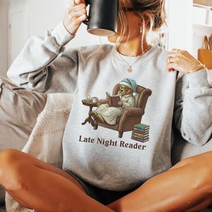 Gildan 18000 sweatshirt featuring a cozy teddy bear wearing a nightgown and hat, sitting in a chair reading a book with a candle, coffee cup, and stack of books beside him. The design includes the text “Late Night Reader” beneath the image. Styled in a warm, vintage-inspired look perfect for book lovers, cozy fall nights, and readers who enjoy a comforting, bookish aesthetic.