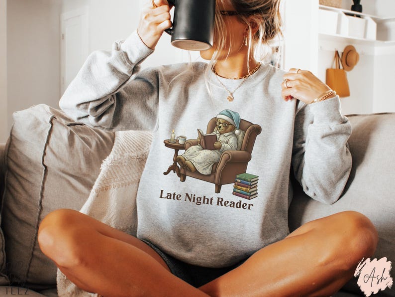 Gildan 18000 sweatshirt featuring a cozy teddy bear wearing a nightgown and hat, sitting in a chair reading a book with a candle, coffee cup, and stack of books beside him. The design includes the text “Late Night Reader” beneath the image. Styled in a warm, vintage-inspired look perfect for book lovers, cozy fall nights, and readers who enjoy a comforting, bookish aesthetic.