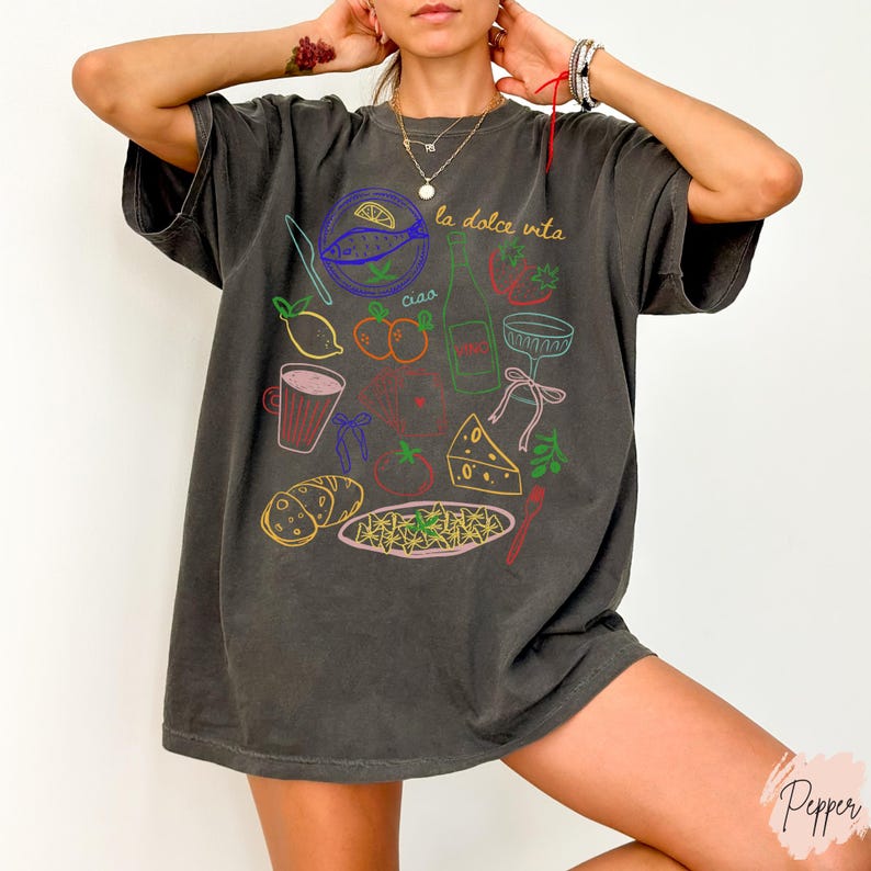 K&ouml;nnte beinhalten: Dunkelgraues T-Shirt mit einer farbenfrohen Grafik italienischer Speisen und Getr&auml;nke, darunter Fisch, Weinflasche, Pasta und K&auml;se. Die Worte "la dolce vita" und "ciao" sind ebenfalls auf dem Shirt zu sehen.